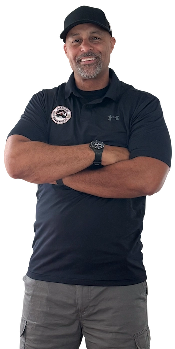 Man standing and smiling, wearing a black cap, black collared Under Armour shirt with a patch, gray pants, and a watch.